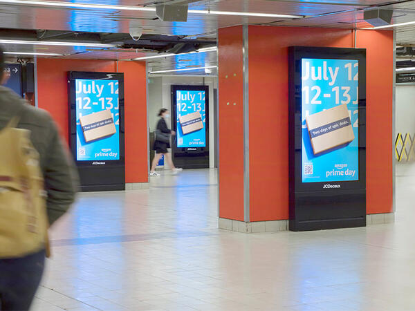 A digital advertising display inside a train station. The screen showcases vibrant and engaging advertisements promoting various products and services. People can be seen passing by.