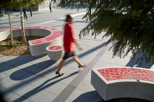 In the early morning light, a person in red walks through the Castle Hill station plaza. To either side of them are trees and round and curved furniture, inlaid with red panels containing tactile drawings and text.