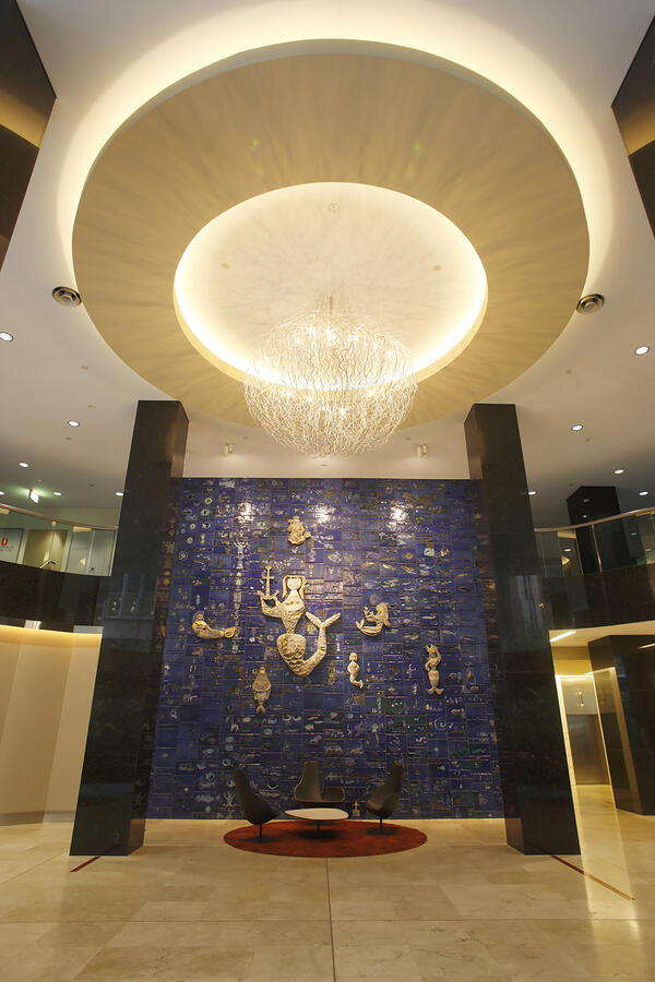 In the foyer of the P&O building, a tiled wall mural is framed between two square black columns. Tiles of navy blue resembling the sea are accented by a large gold swimming mermaid, surrounded by six smaller mermaids.