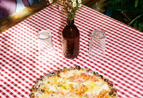Pizza sits on table covered in red and white check cloth, two glasses and vase