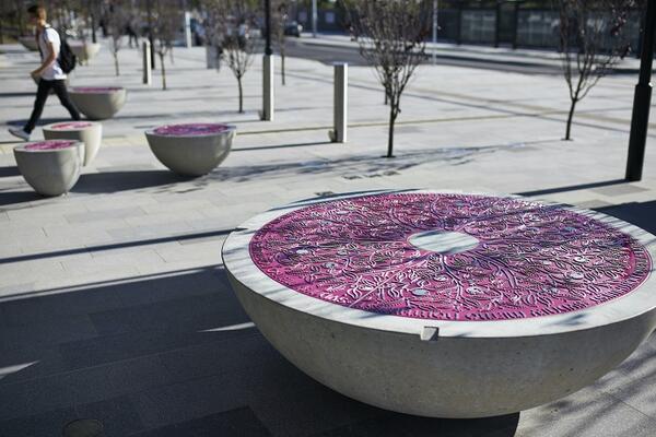 A large circular seat is placed in a paved plaza, rounded at the bottom like a piece of fruit cut in half. On its top, a blossom pink panel contains detailed tactile drawings of leaves and flowers. In the background, more seats in different sizes are seen.