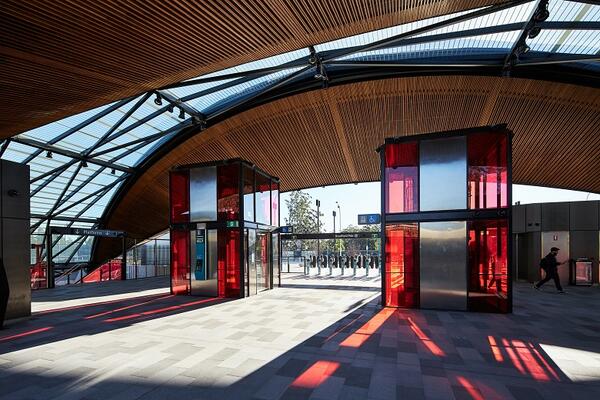 The sun shines behind the concourse of Cherrybrook station, framed by a curved timber roofs. The cherry red glass of the two lifts casts long bright shadows on the floor of the station.