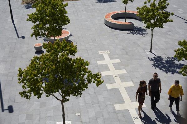 From above, the plaza of Hills Showground station is pictured with three people walking through it. Around them are rows of trees surrounded by rounded furniture topped by bright orange panels. A hopscotch tile runs through the rows of trees.