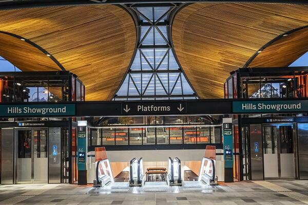 The entrance to Hills Showground station has two central escalators and a lift to each side, all with deep orange glass.A curved roof above frames early evening sky, and skylight lanterns in the background glow with orange colour.