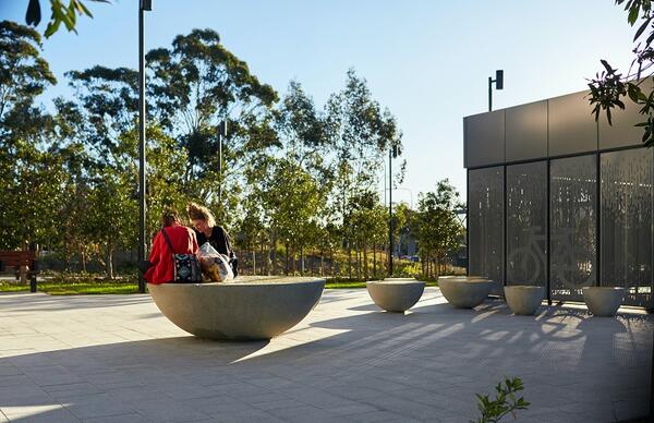 In the plaza of Kellyville station, morning light frames a grove of trees, and round seating elements. Two people are sitting on one of the seats, which are topped by inlaid yellow discs.