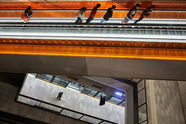 From above, a collection of customers are seen riding up the escalator at Norwest station. Below them is the station platform. Light orange glass highlights the escalator against the grey of the station flooring.