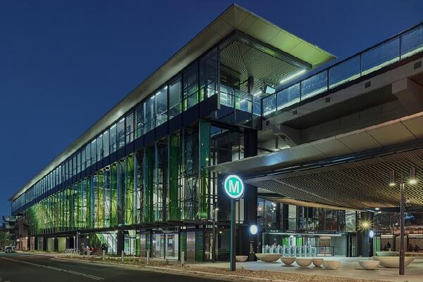 Rouse Hill station’s large glass façade is accented with shades of apple green. In the plaza, a teal Metro M sign stands near a collection of round seating. The station glows with colour in the evening sky. 